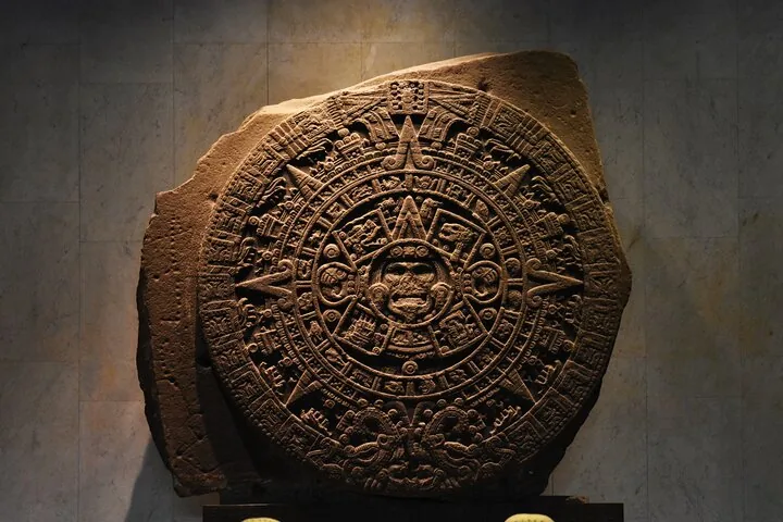 The Aztec Sun Stone displayed against a plain wall in a museum.